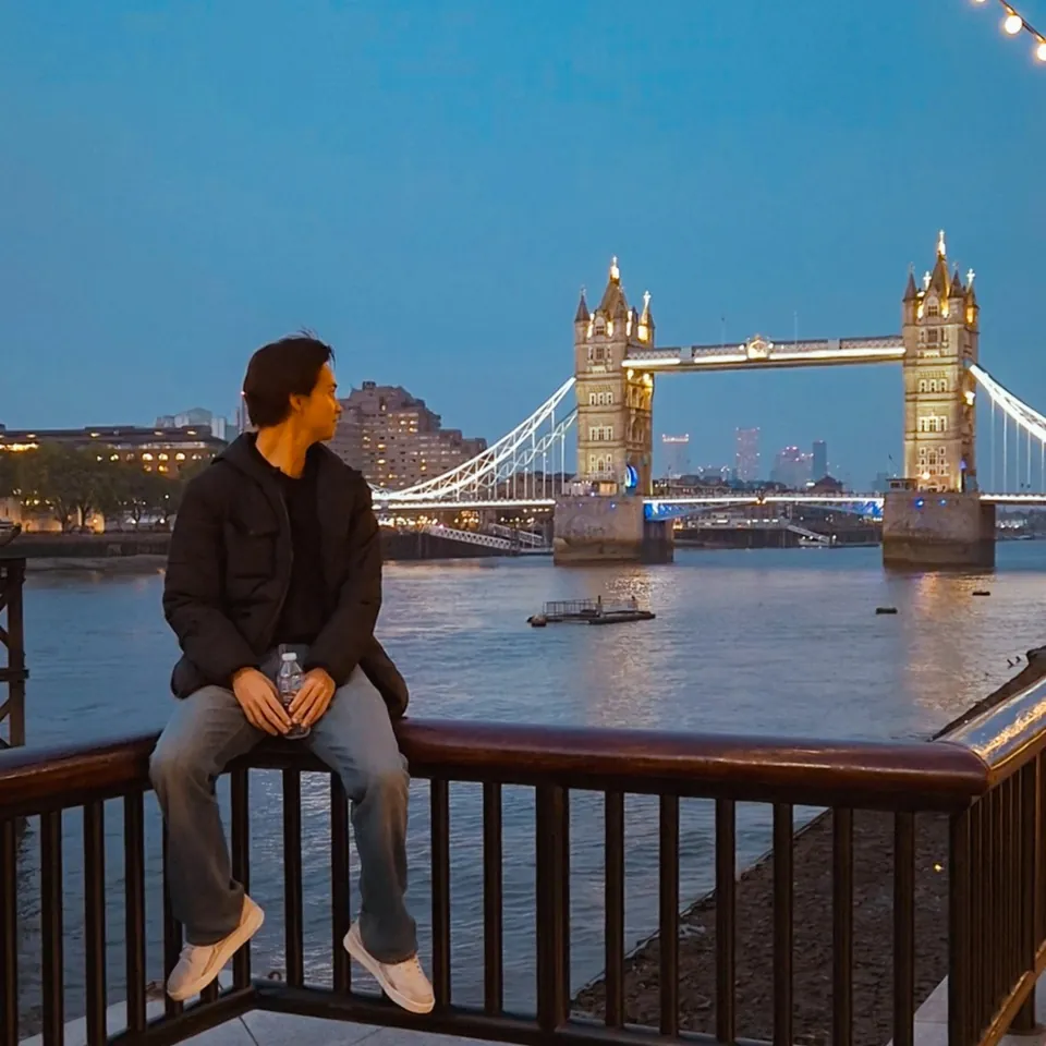 Joven sentado en una baranda frente al Tower Bridge de Londres al atardecer