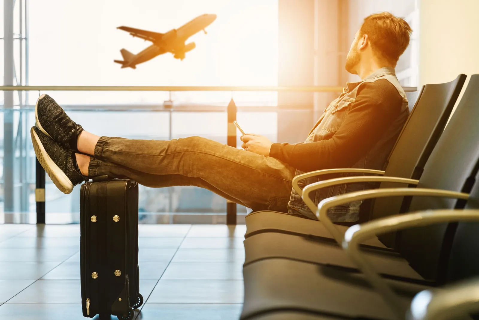 Joven esperando su vuelo en un aeropuerto, con la maleta al lado